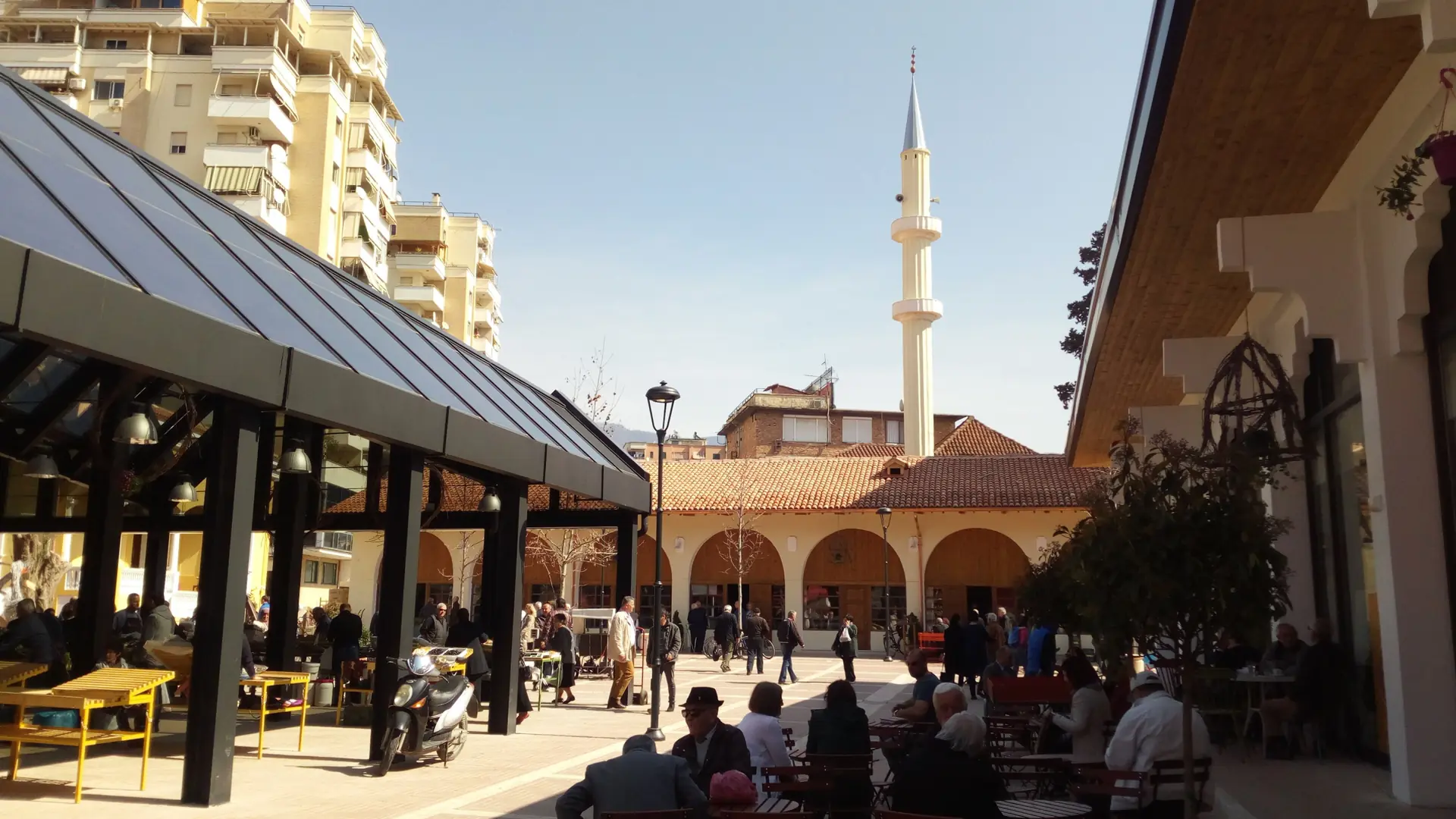 Café tables under the canopies of the New Bazaar (Pazari i Ri) in Tirana, with a minaret beyond, on a sunny day