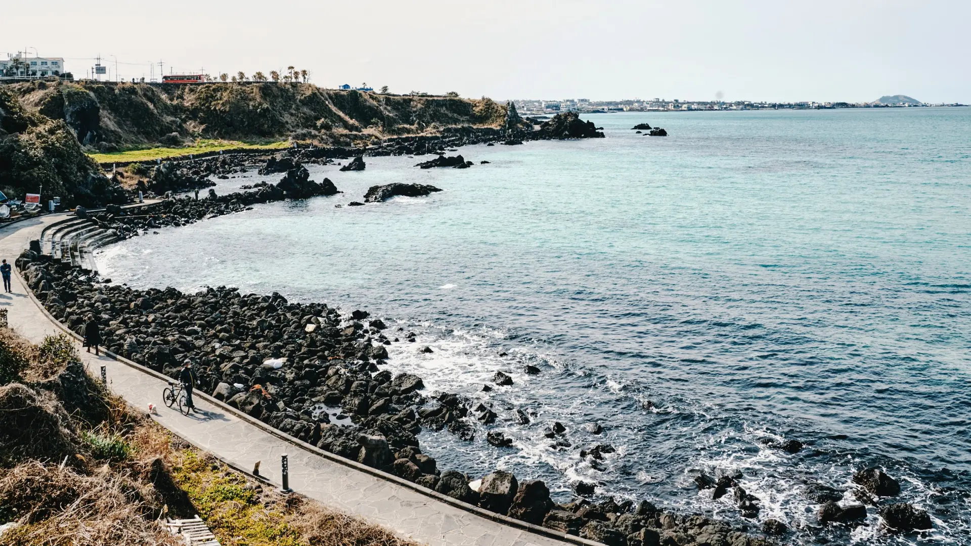 The Handam coastal path curves beside black volcanic rocks and blue water in Aewol