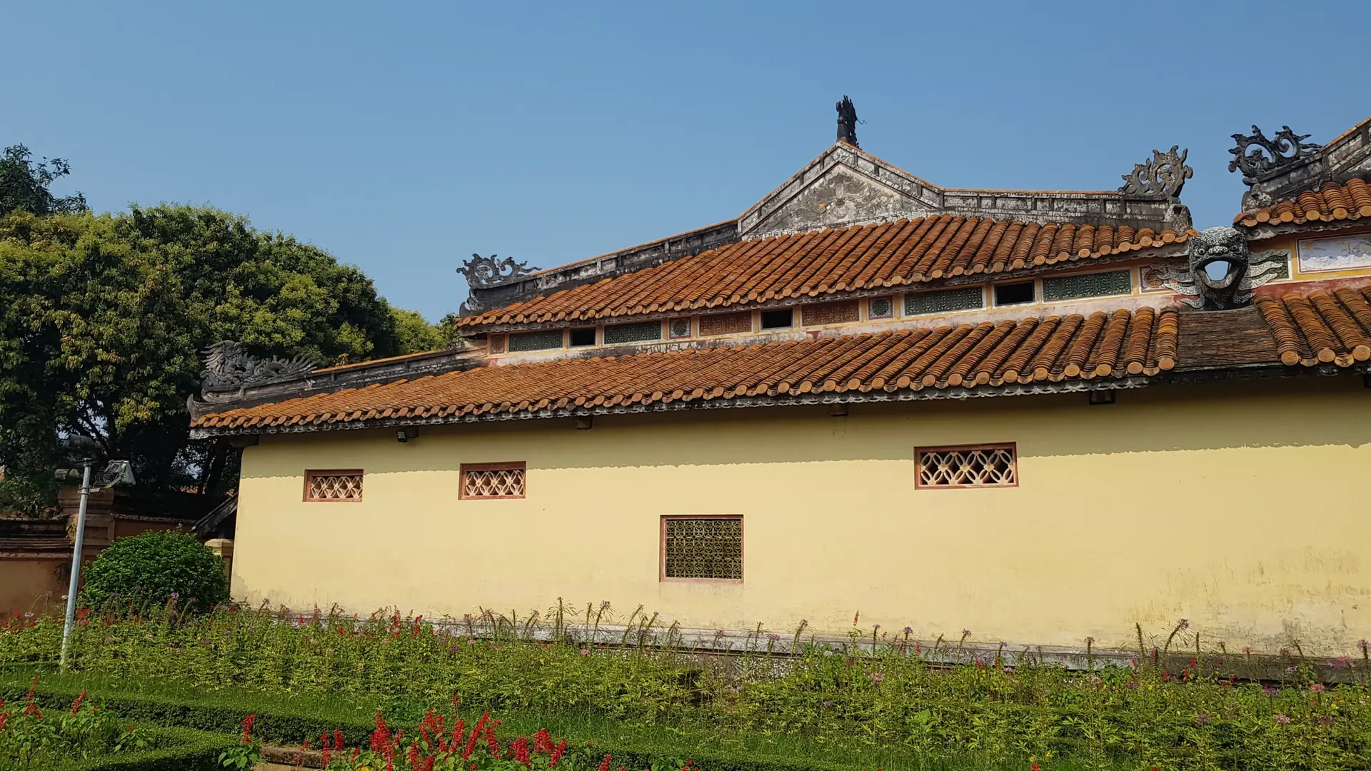 Sunlit tiled roofs, a yellow heritage wall and flowers in wider Hue