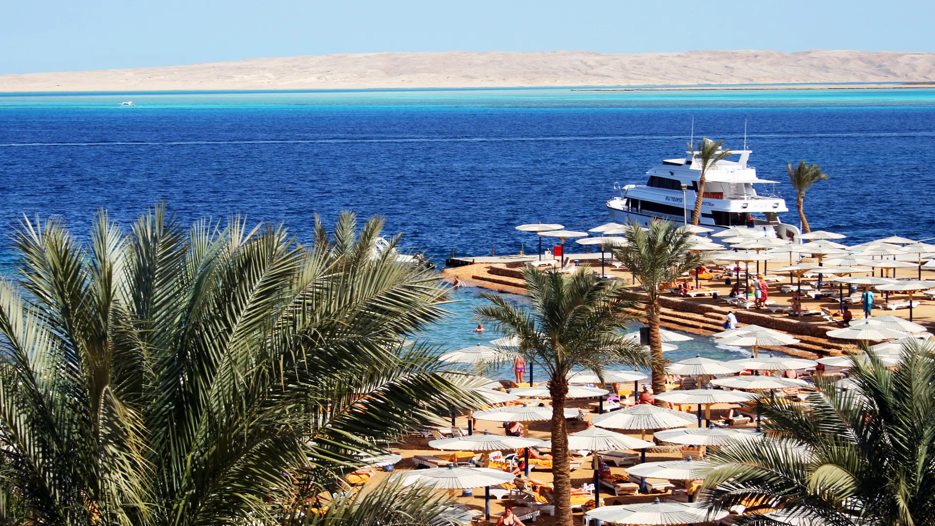 Palm-lined Hurghada hotel beach and marina beside deep blue Red Sea water in bright sun.