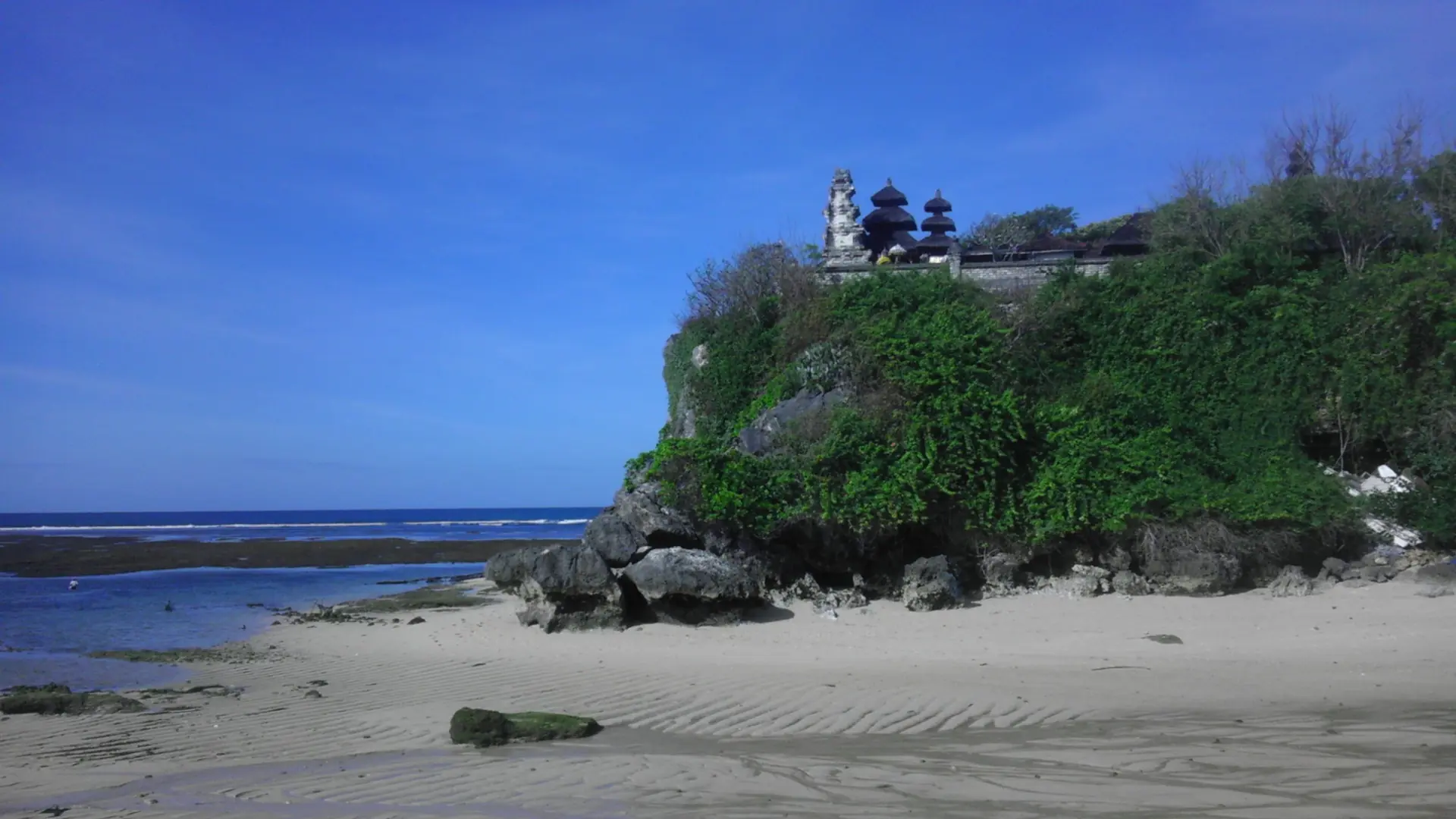 Pura Geger’s tiered temple roofs above a green rocky headland and pale beach in Nusa Dua, Bali