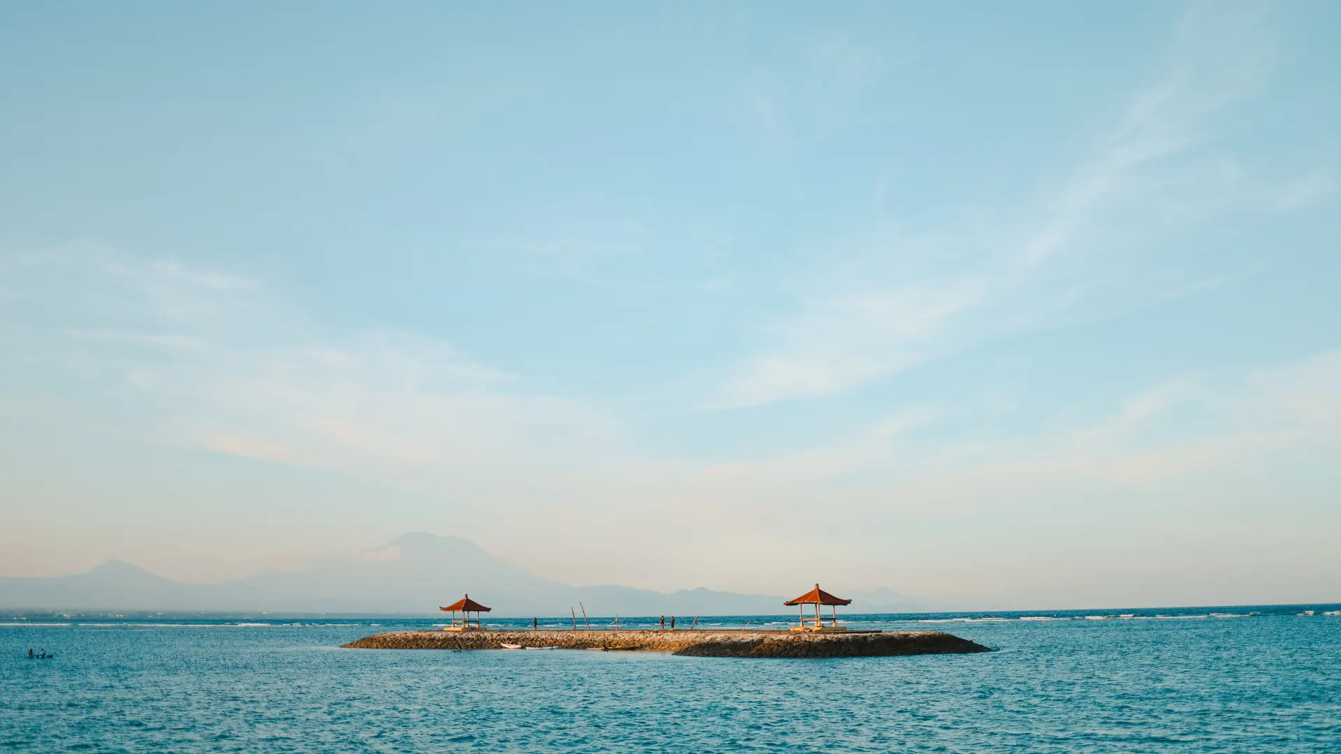 Two red-roofed pavilions on a rocky breakwater in Sanur’s blue water, with Mount Agung in the distance