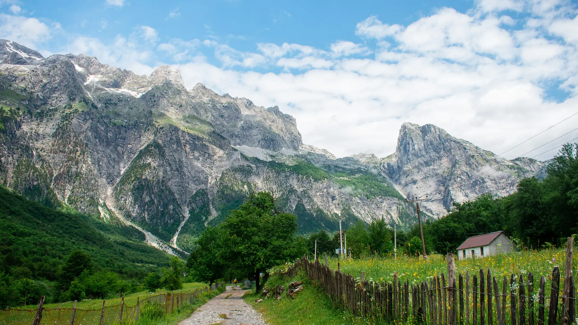 A farm track and wooden fences leading into the Theth valley below limestone peaks on a clear summer day