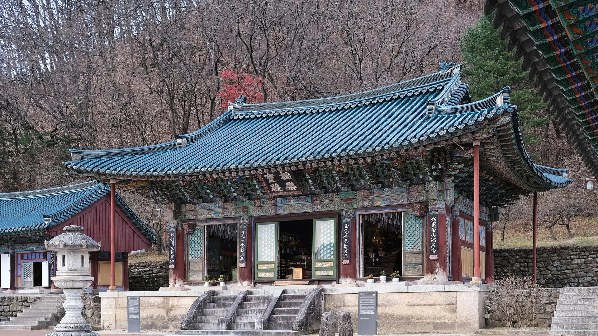 A painted temple hall with a blue tiled roof at Sinheungsa, with steps and bare trees on the hillside behind