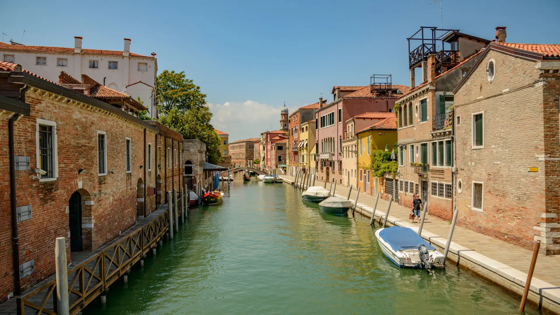 a sunlit Venetian canal with a low stone bridge, terracotta façades and moored boats