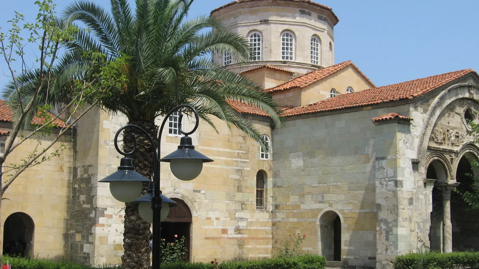 Hagia Sophia of Trabzon behind a palm and curved garden lamps, shown as a wider city scene