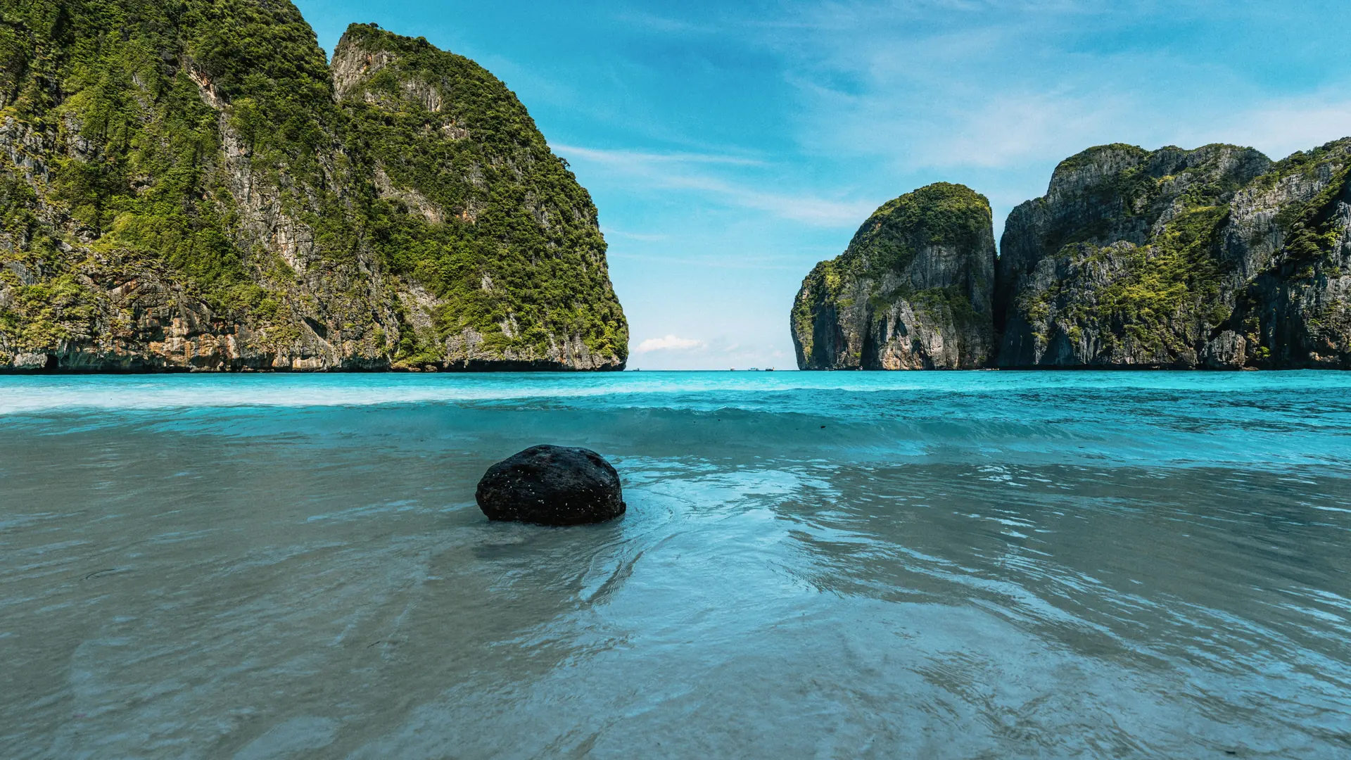Sunlit limestone walls and turquoise shallows around a dark rock at Maya Bay, Phi-Phi Leh