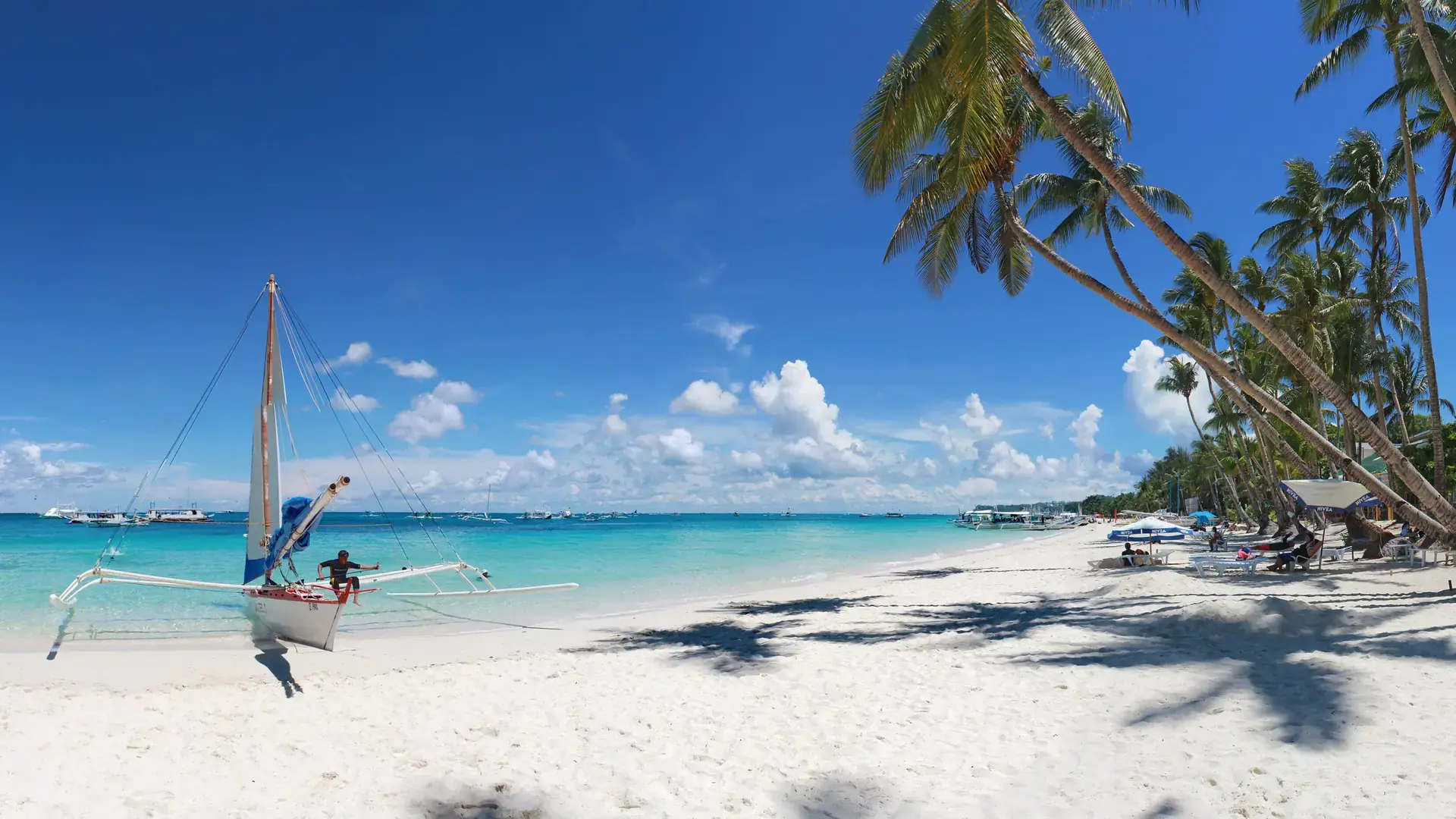 A paraw sailboat beside the white sand and palms of Boracay’s White Beach under a blue sky
