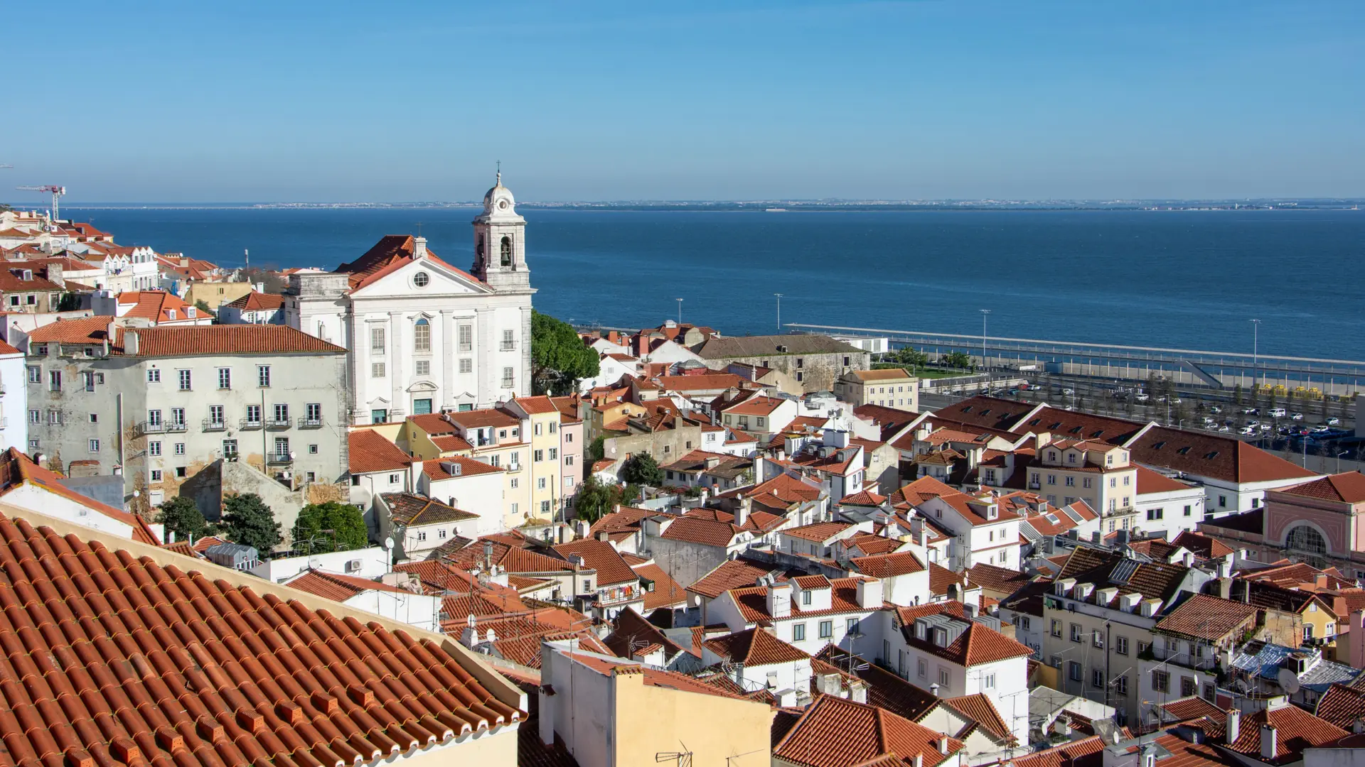 Lisbon’s terracotta rooftops and a white church overlooking the blue Tagus River in daylight