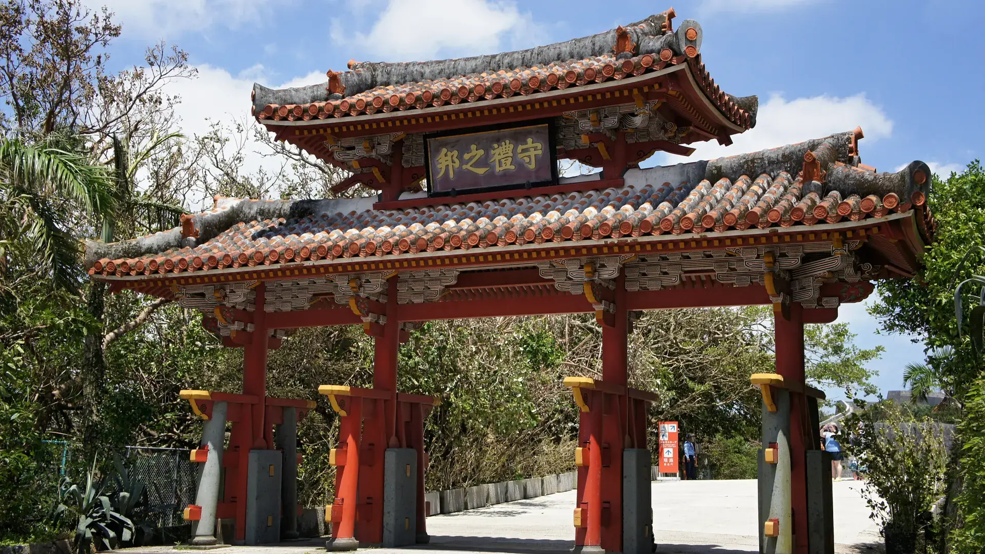 Shureimon's vermilion gate at Shurijo Castle Park framed by tropical green trees and blue sky