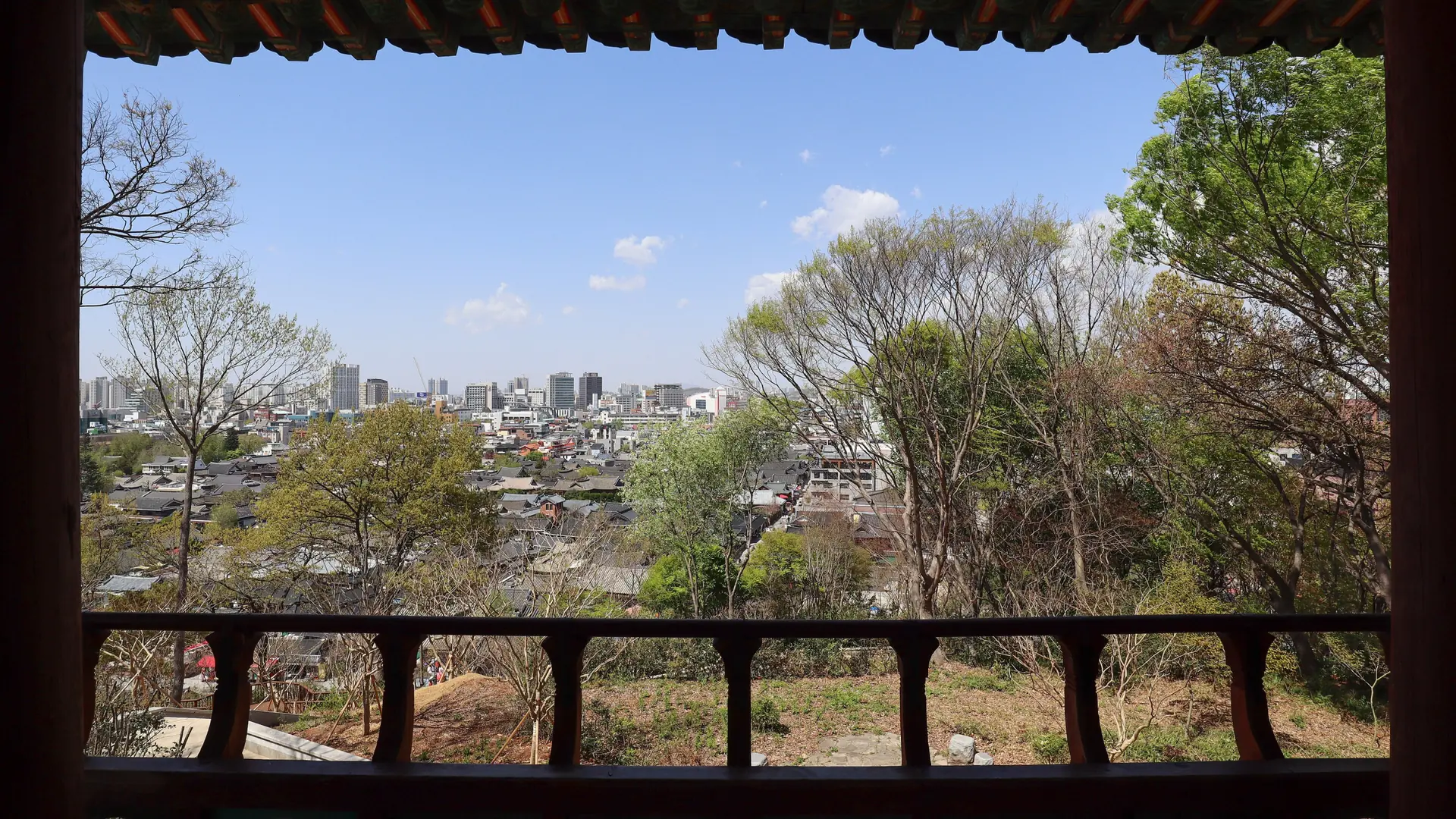 View from Omokdae pavilion across trees and Jeonju rooftops, framed by a wooden railing and tiled eaves