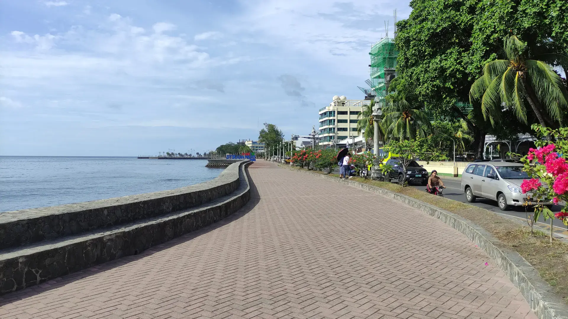 Rizal Boulevard’s paved waterfront walk beside blue water, flowering shrubs and city buildings in Dumaguete