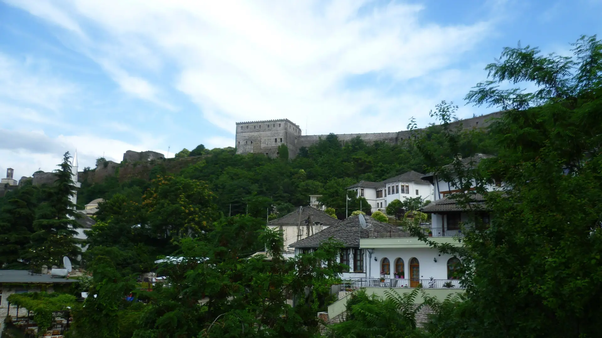 Old stone houses among trees below the walls of Gjirokastër castle on a clear day