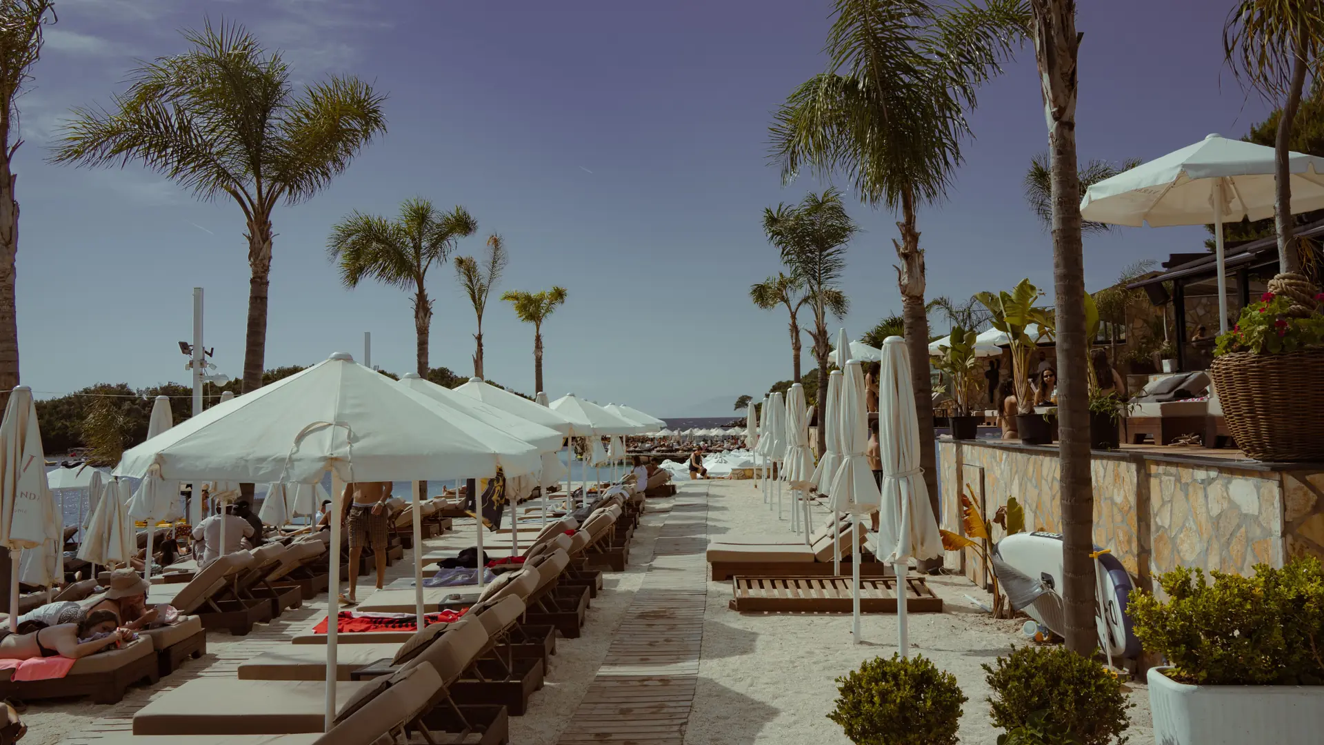 Palm trees, white umbrellas and a beach-club deck along the waterfront at Ksamil on a sunny day