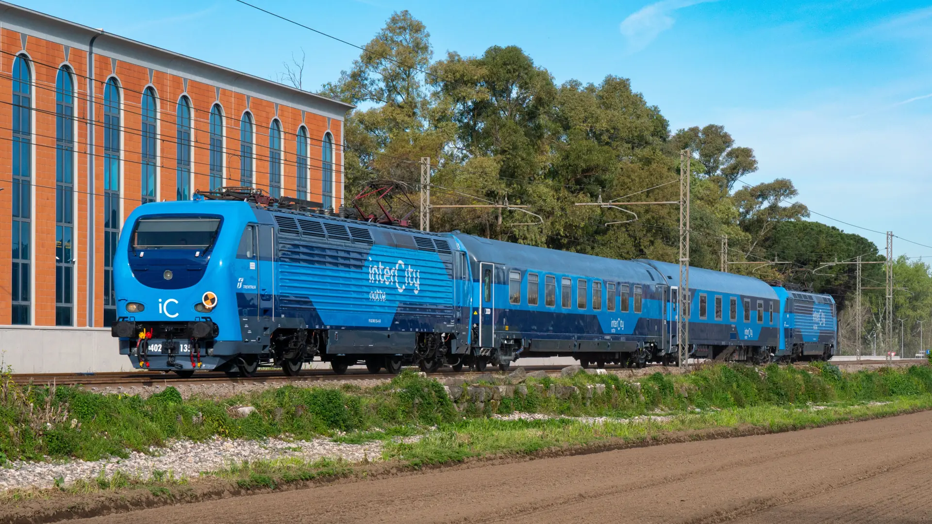 a blue CP Intercity train under a bright blue sky beside green trees