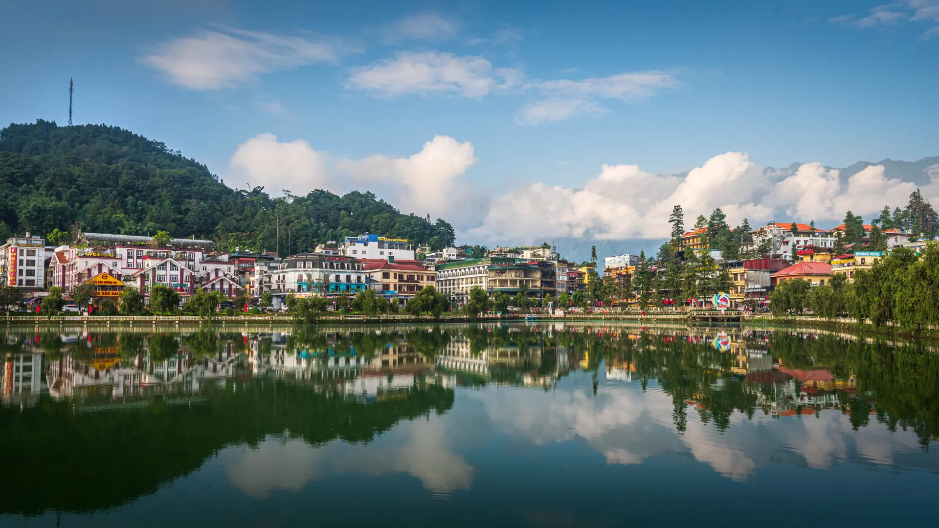 Sunlit Sapa town buildings reflected in the lake below a wooded hillside