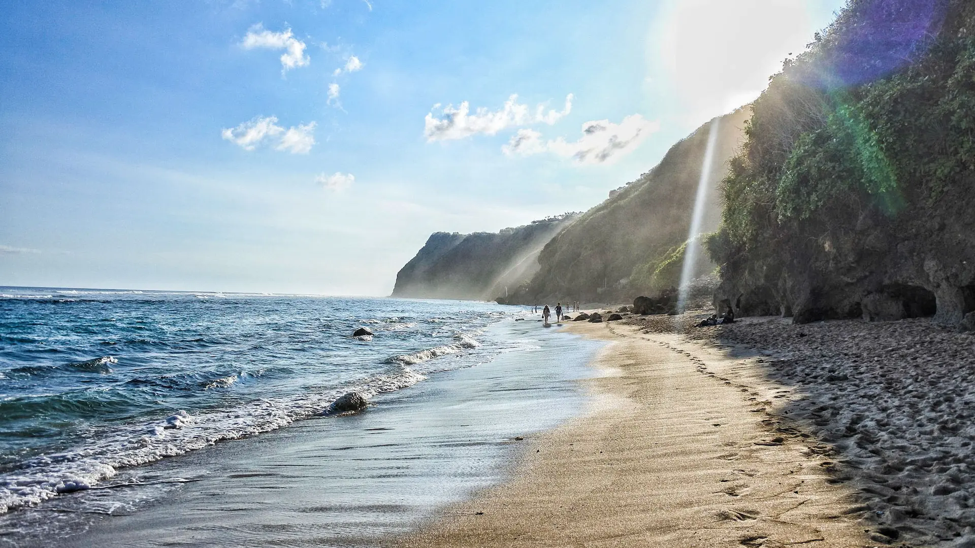 Sunlit wet sand and blue water beside the rocky wider Nusa Dua shoreline in Bali