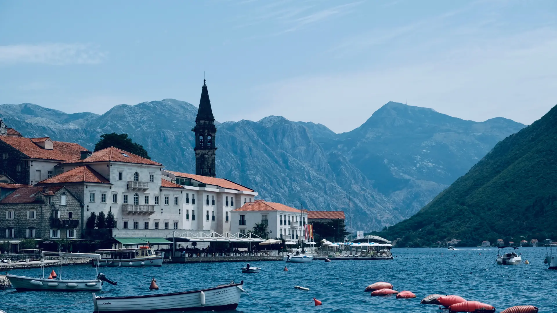 The waterfront of Perast with stone palazzi and a bell tower beside the calm bay on a sunny day