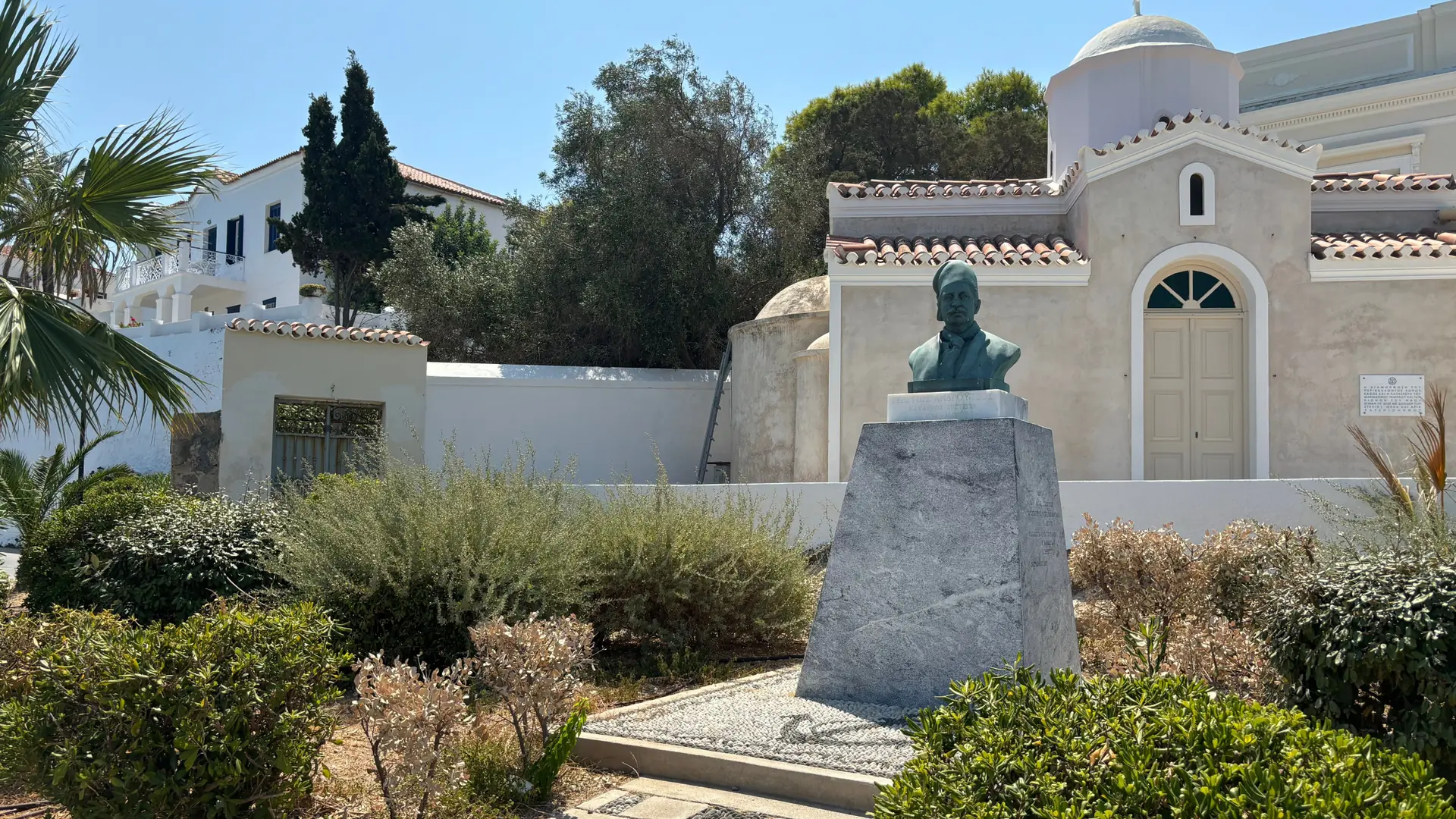 A bronze memorial and white chapel surrounded by palms and gardens on Spetses