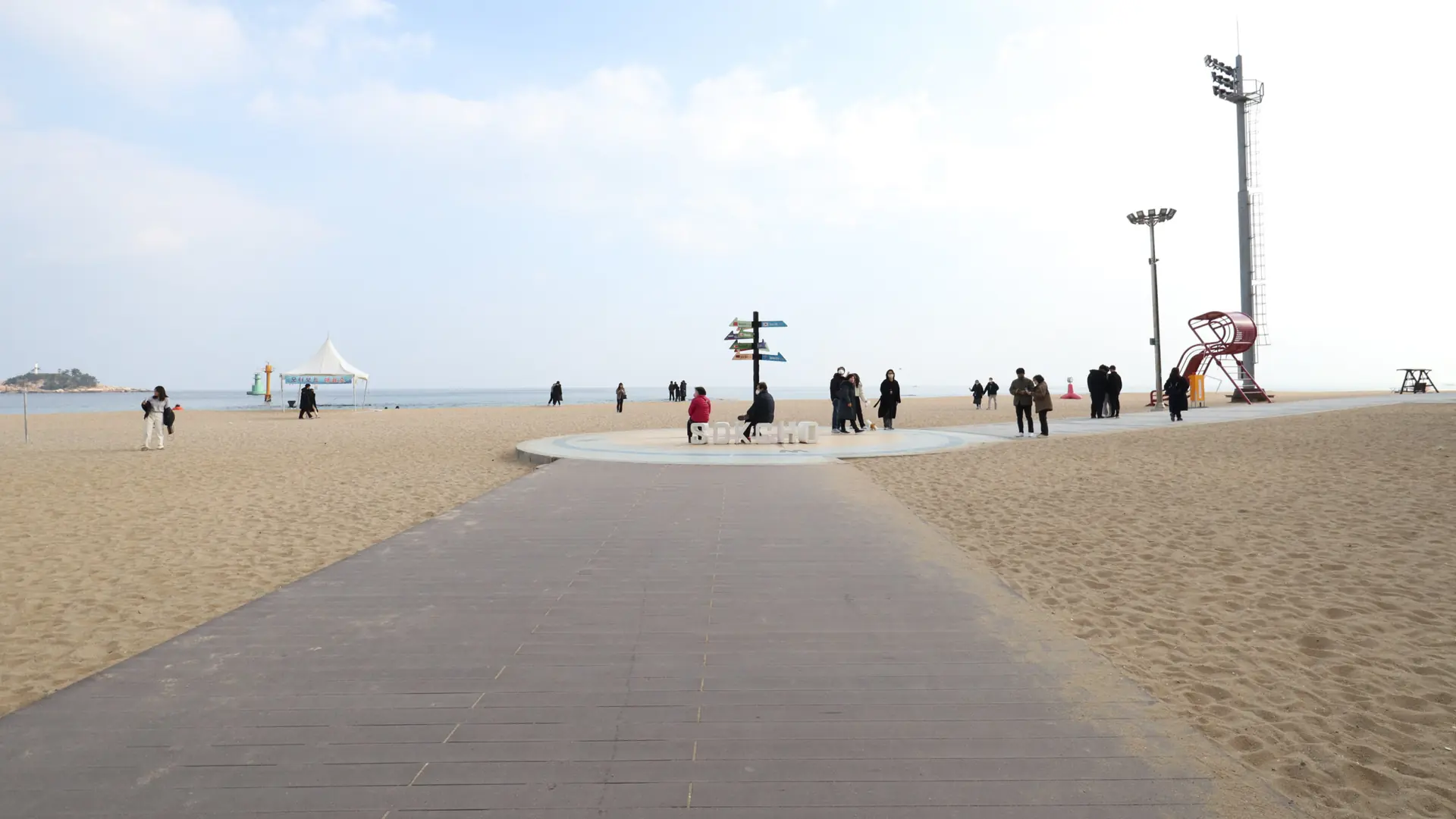 A paved path between sandy sections of Sokcho Beach, with people walking towards the sea