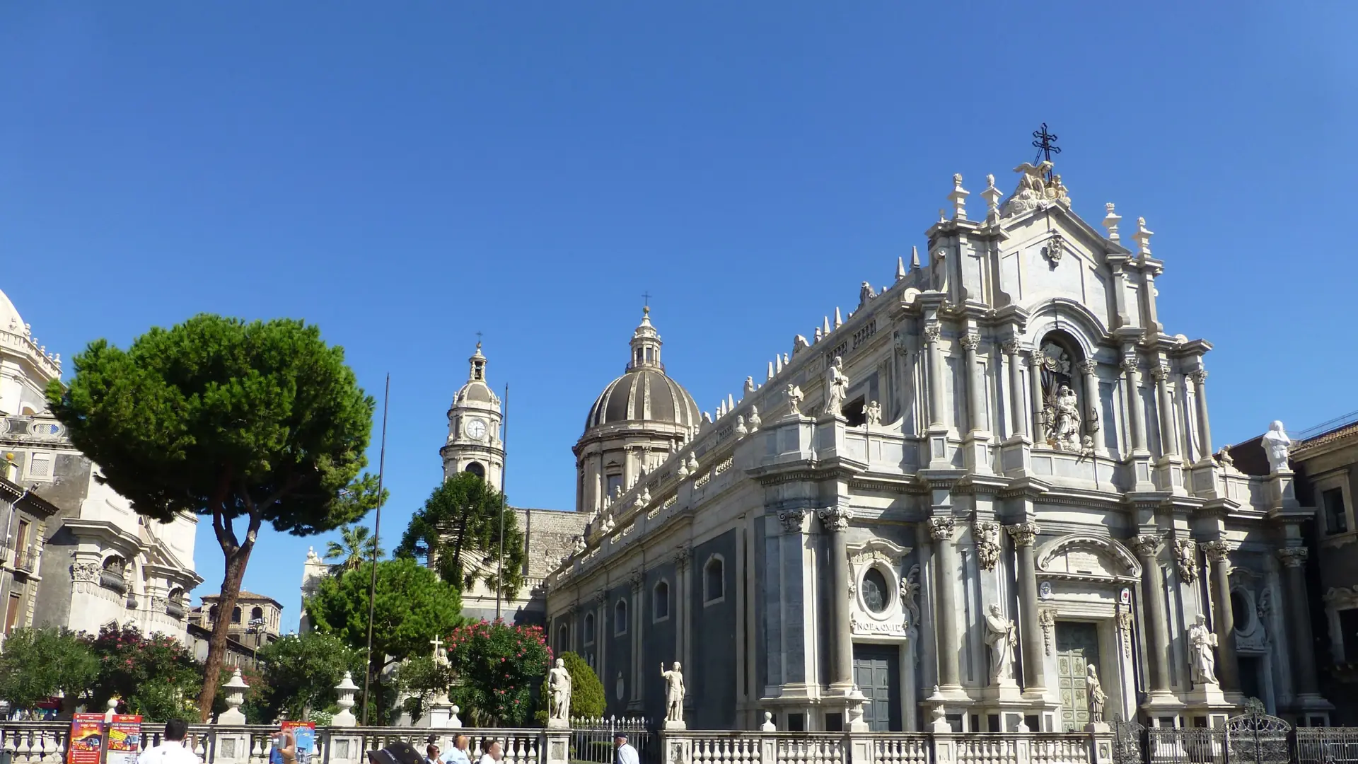 Catania Cathedral's Baroque limestone facade under a clear blue sky, framed by pine trees
