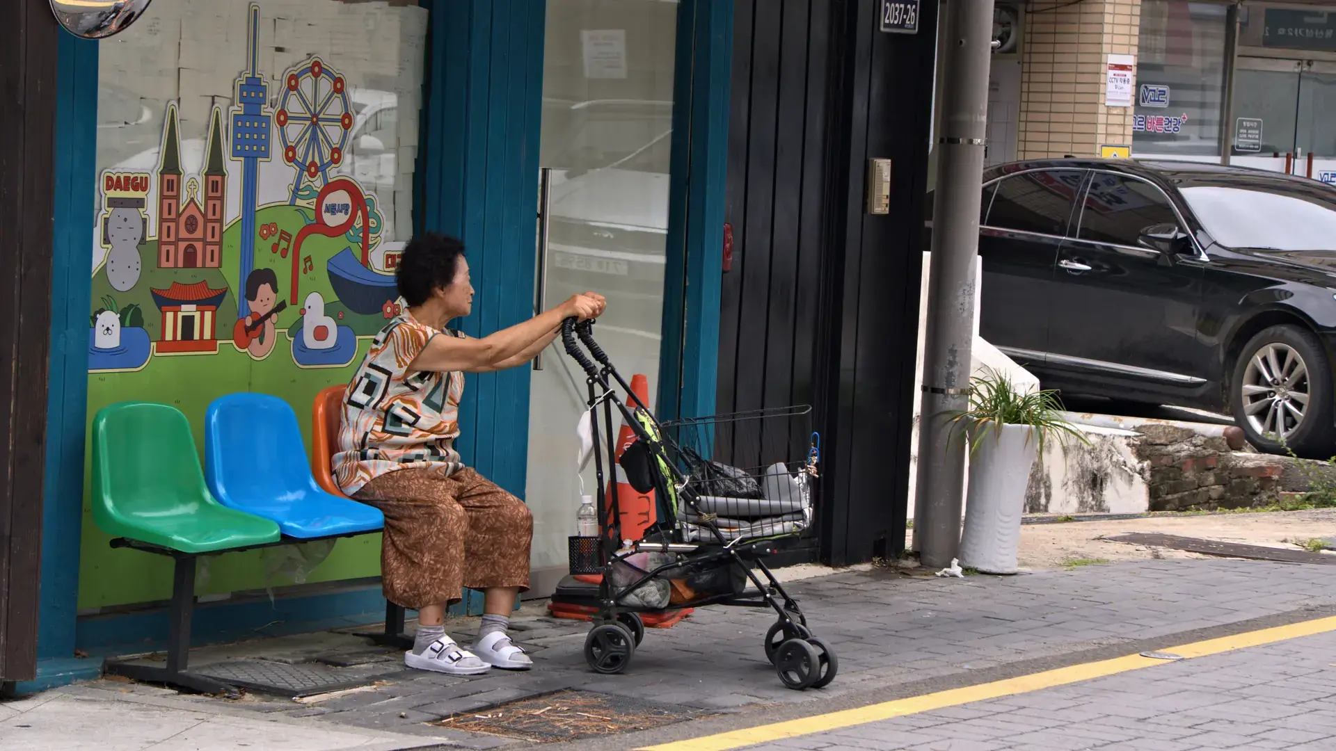 A woman waiting at a bus stop painted with a Daegu city mural, on a sunny street in Daegu, South Korea