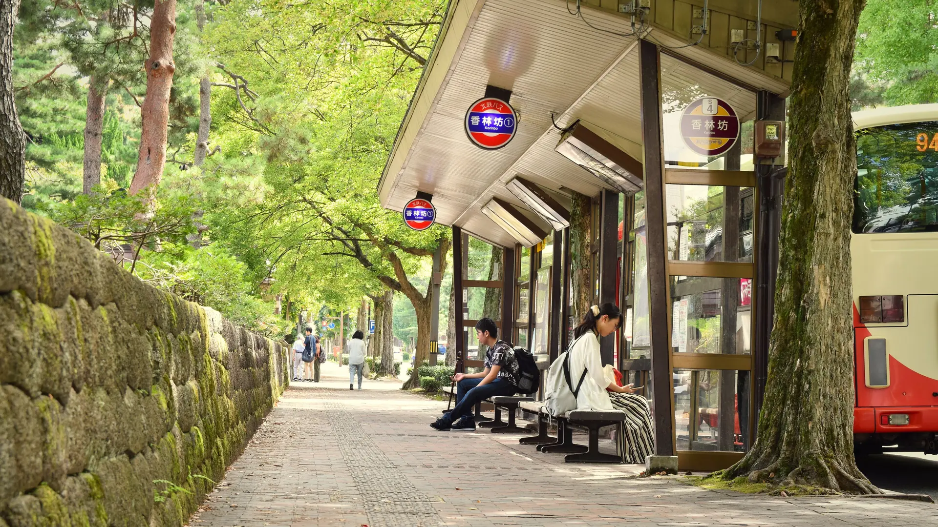 Sunlit trees, stone walls and benches beside Korinbo’s signed bus stop in Kanazawa