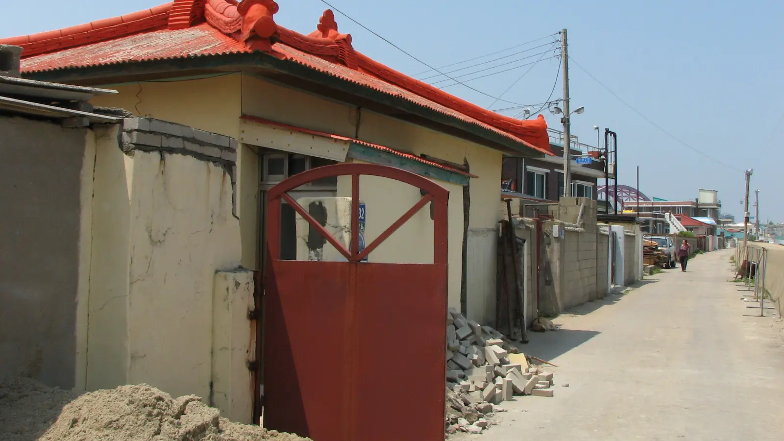 A narrow Abai Village lane past a house with a red-tiled roof and gate, under clear blue sky