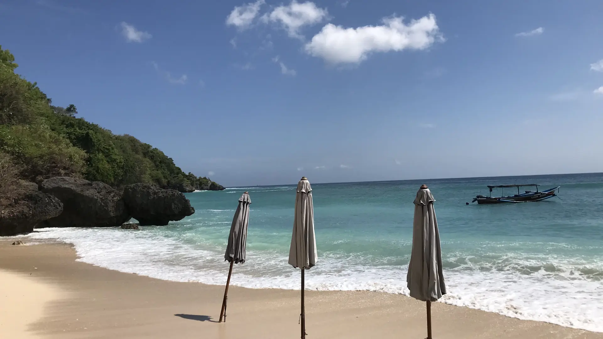 Pale sand, closed beach umbrellas and a traditional boat beside limestone formations at Padang Padang beach in Bali