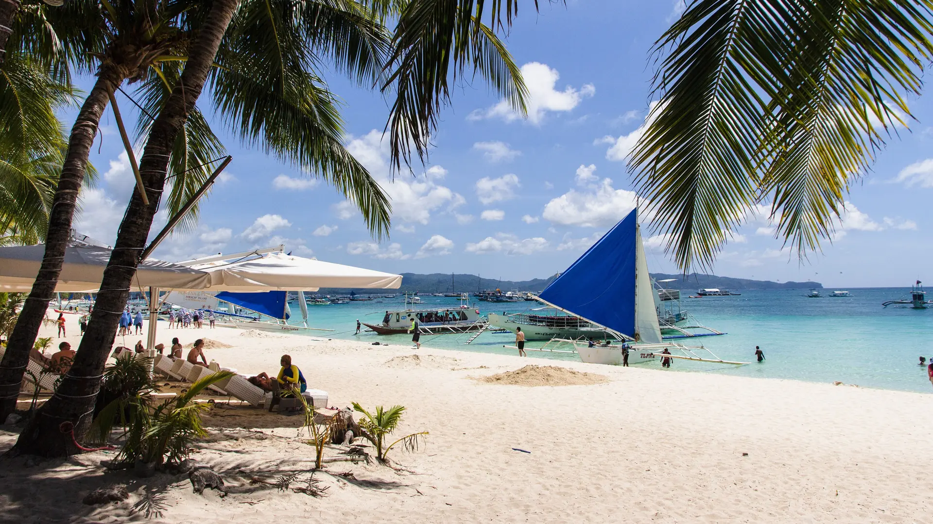 Palm-framed White Beach on Boracay, with a blue-sailed paraw, beachgoers and turquoise water