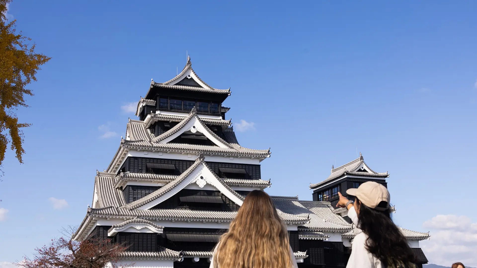 Kumamoto Castle’s black-and-white keeps beneath a blue sky, with two visitors in the foreground.