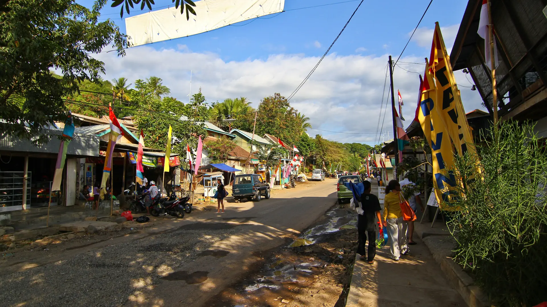 Shops, pedestrians and colourful flags along Labuan Bajo’s main street in daylight