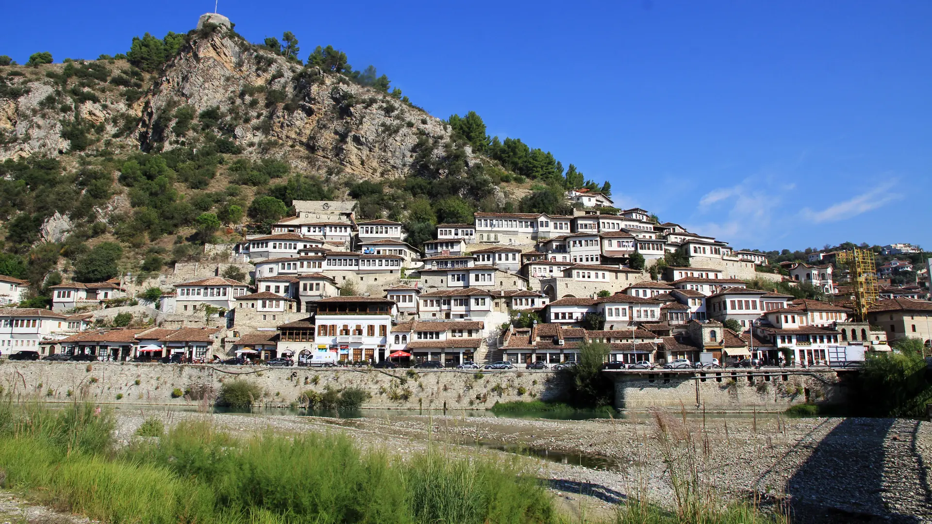 The white Ottoman houses of Berat's Mangalem quarter climbing the hillside below the castle, above the Osum river on a sunny day