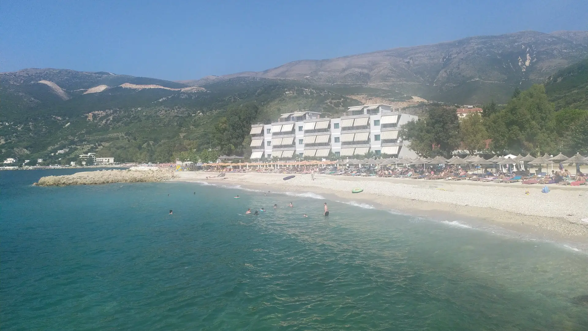 Turquoise water and a curving beach backed by hotels at Radhimë, south of Vlorë, under a bright sky