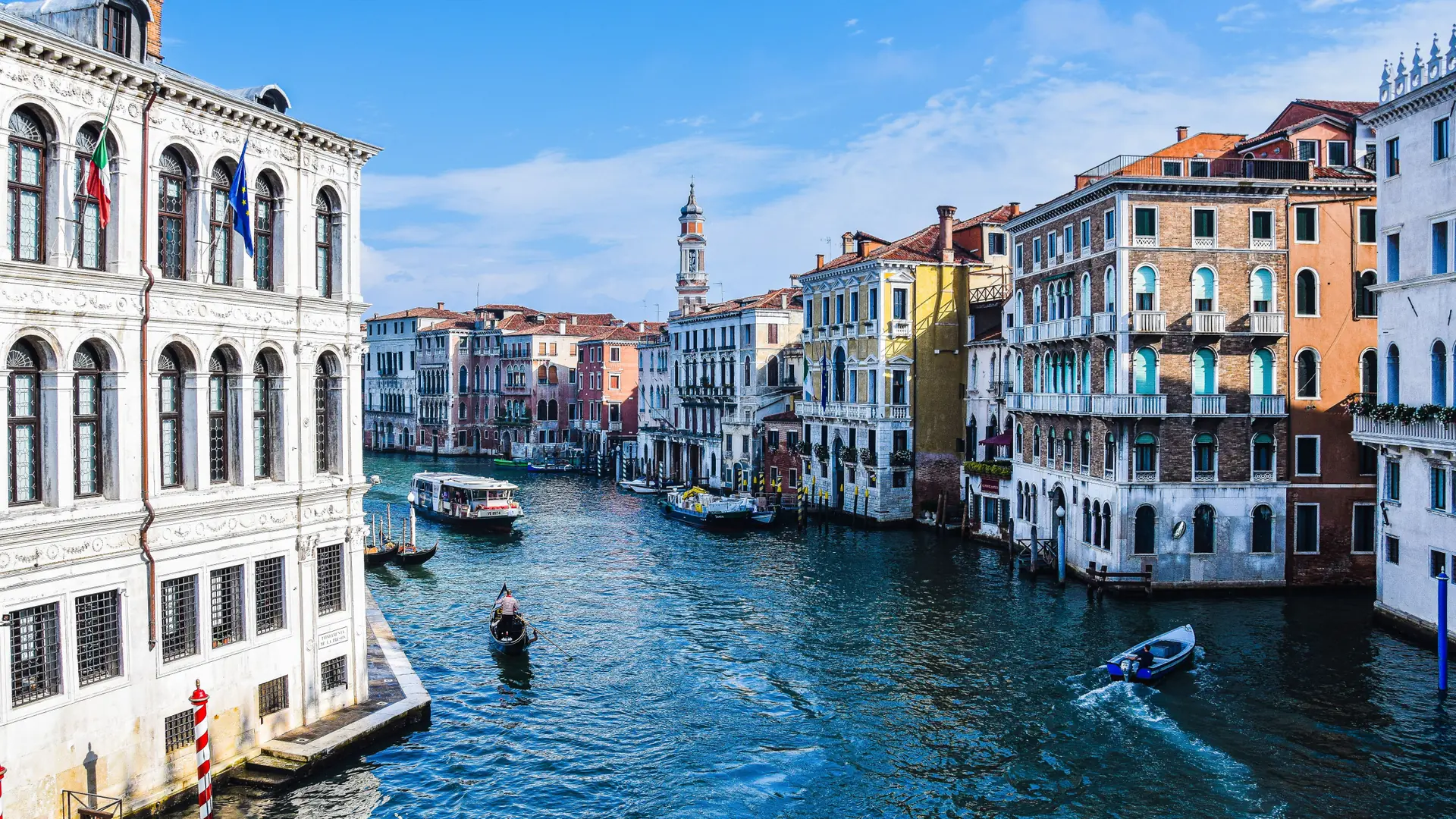Boats on Venice’s Grand Canal between historic palazzi beneath a bright blue sky.