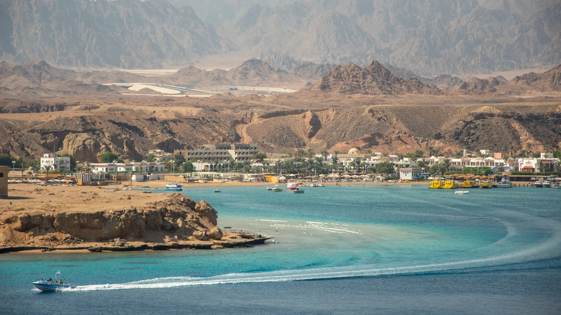 Daylight view across a Sharm el-Sheikh bay, with coastal buildings, a passing boat and desert mountains.