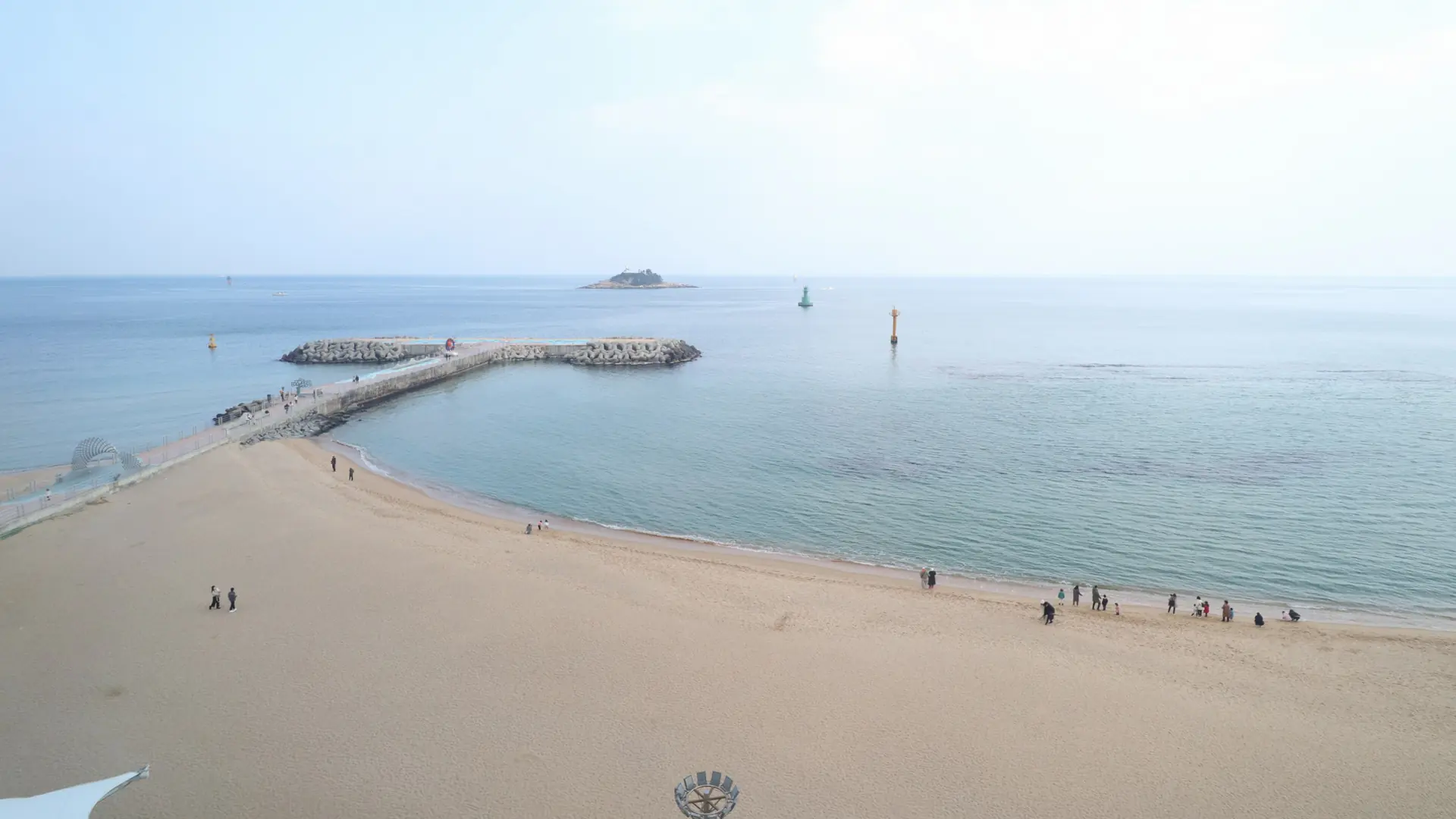 Sokcho Beach viewed from above, with pale sand, people near the water and a curved stone breakwater