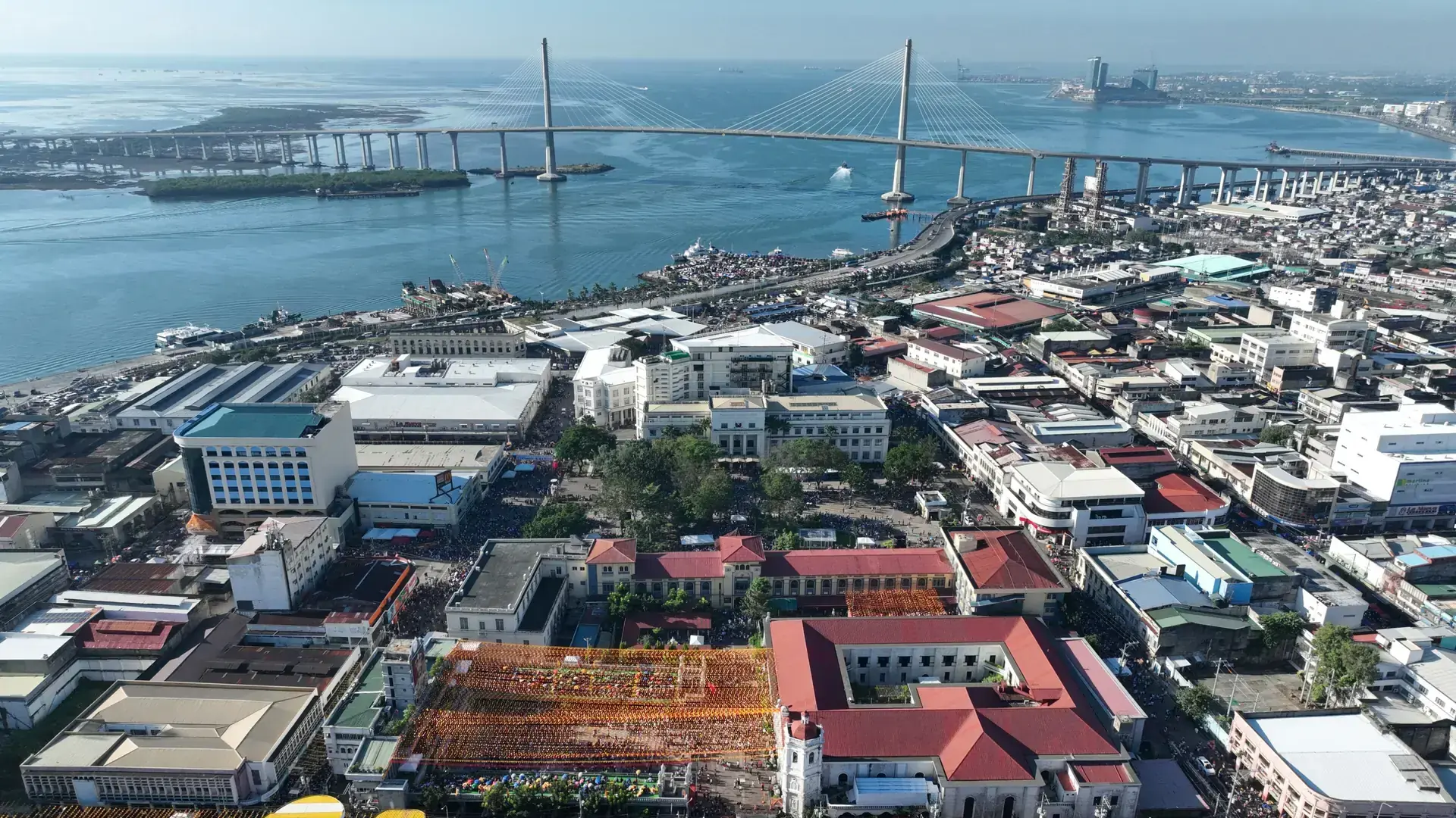 Sunlit downtown Cebu rooftops and the Basilica del Santo Niño, with the CCLEX bridge beyond the harbour