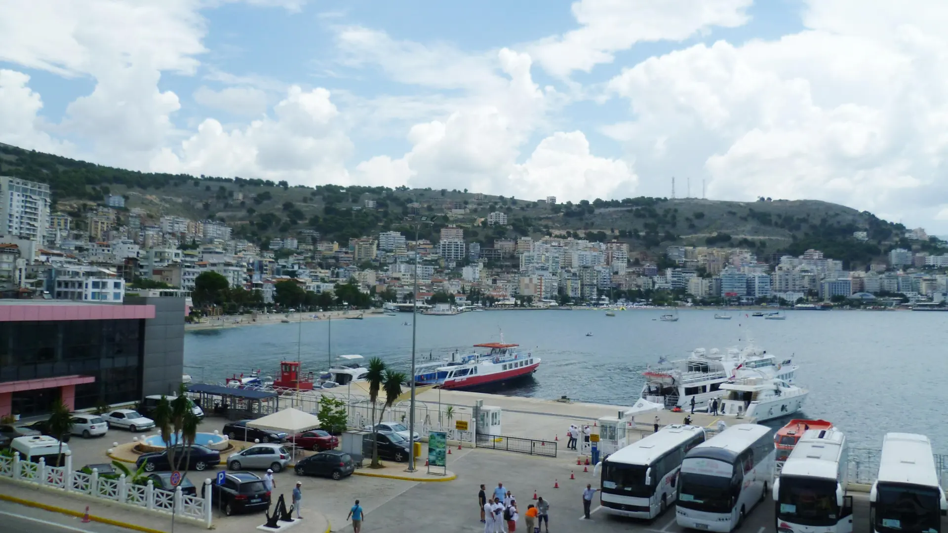 Ferries and coaches at Sarandë's port, with the town climbing the hill behind on a sunny day